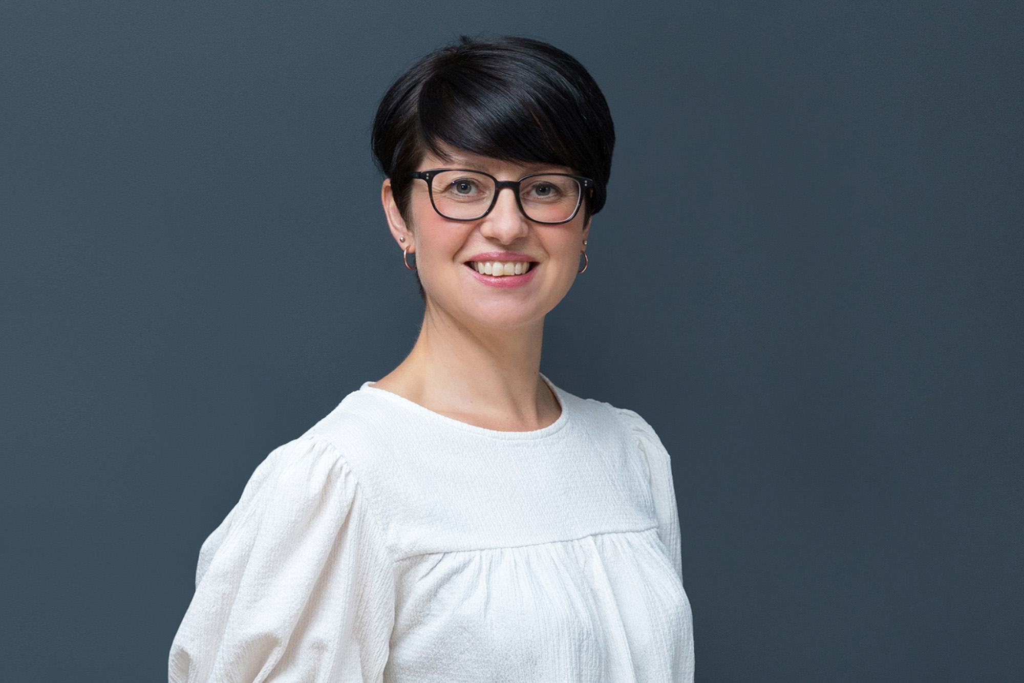 A corporate headshot of a business woman with glasses against a dark grey background; an example of London headshot photography