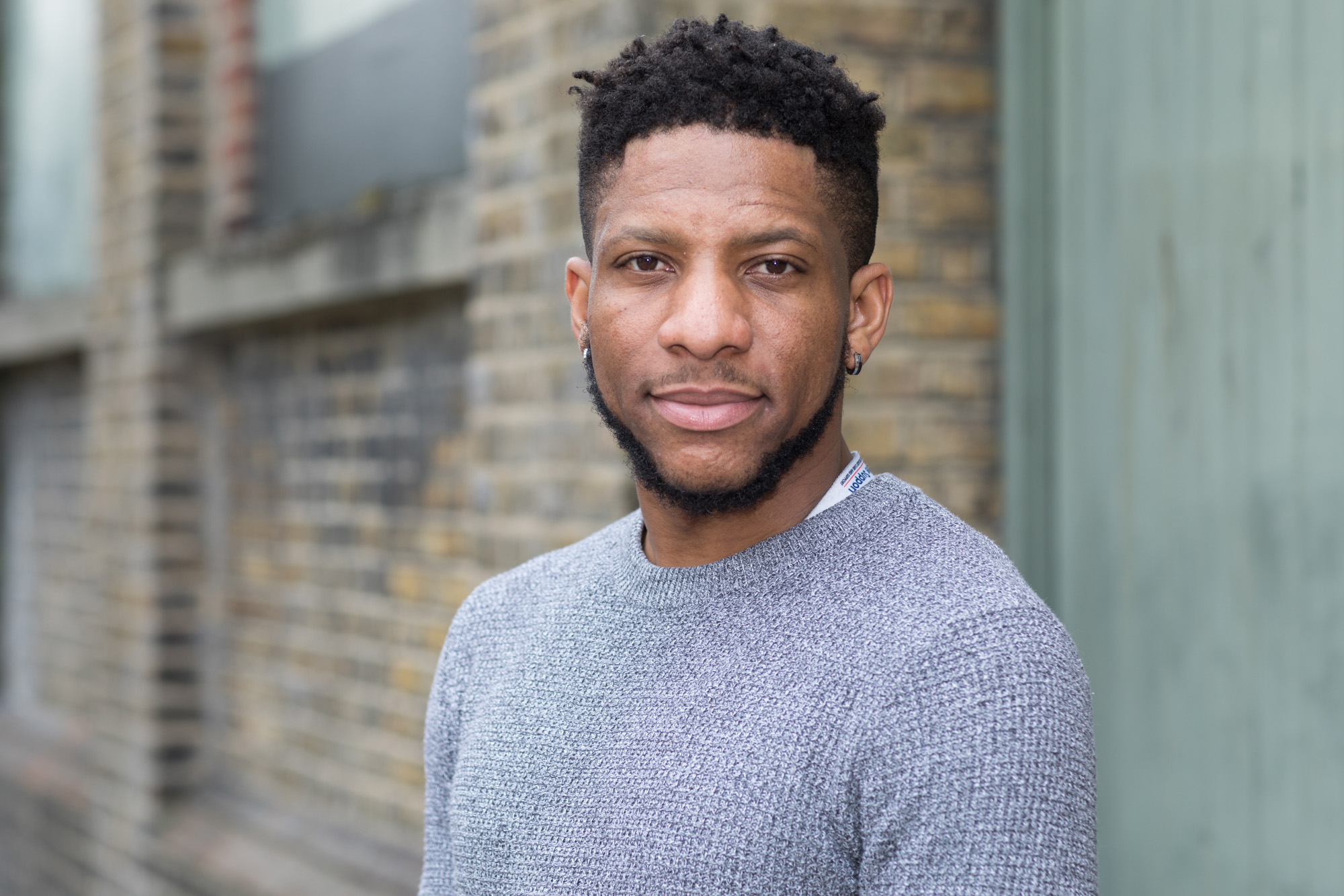 A man ina grey jumper stands in front of a brick wall in Croydon, South London. An example of relaxed headshot photography.
