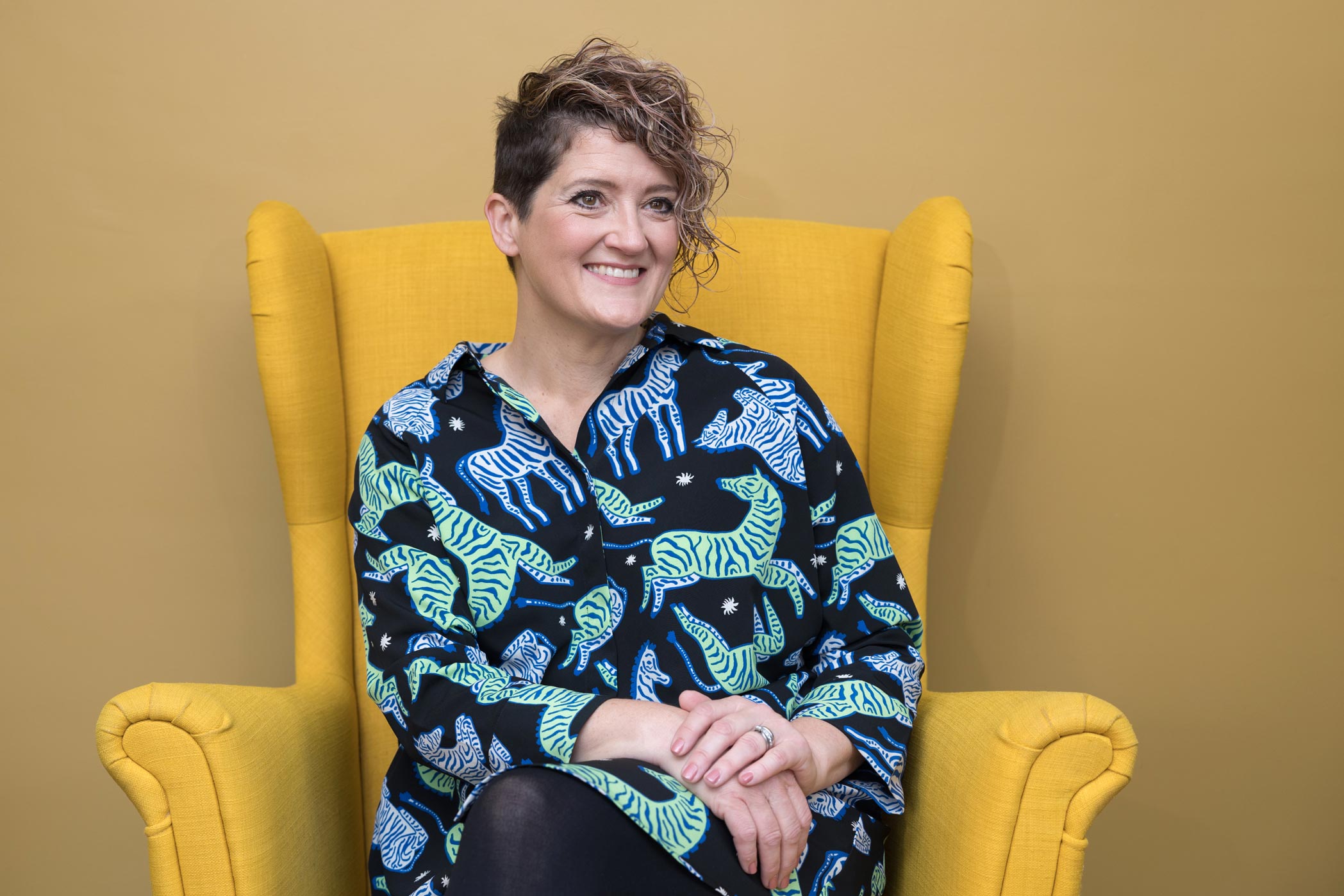 A smiling woman on a yellow chair in front of a yellow wall during her Croydon personal branding photography shoot