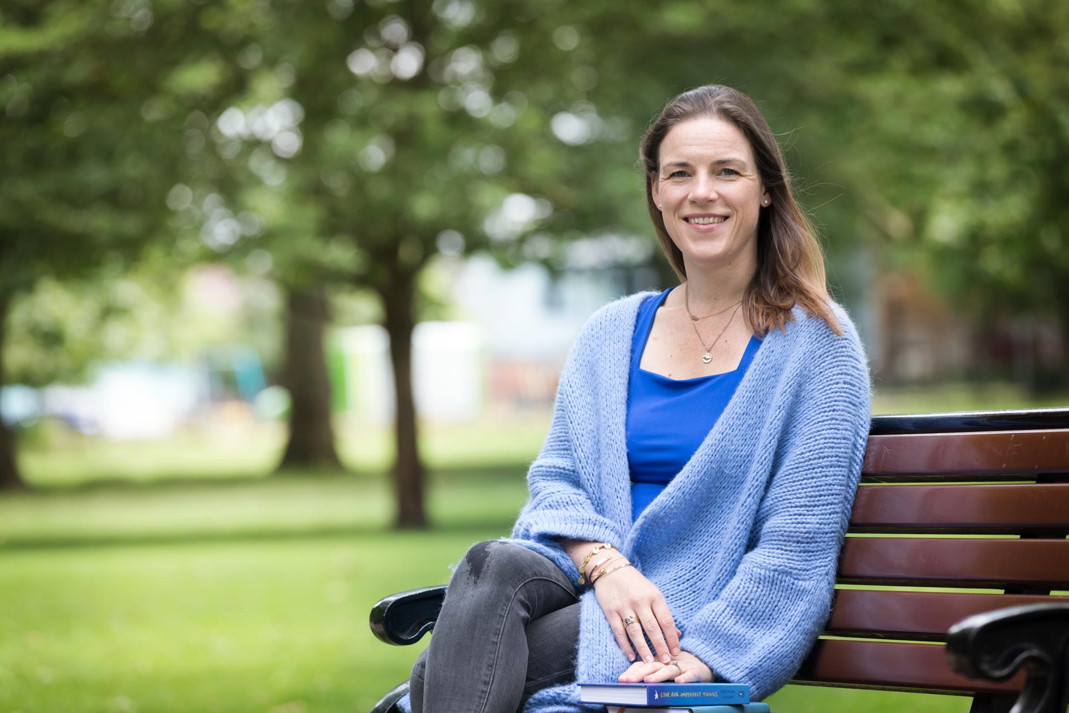 A woman in a blue cardigan sits on a bench in a London park during her personal branding photography shoot