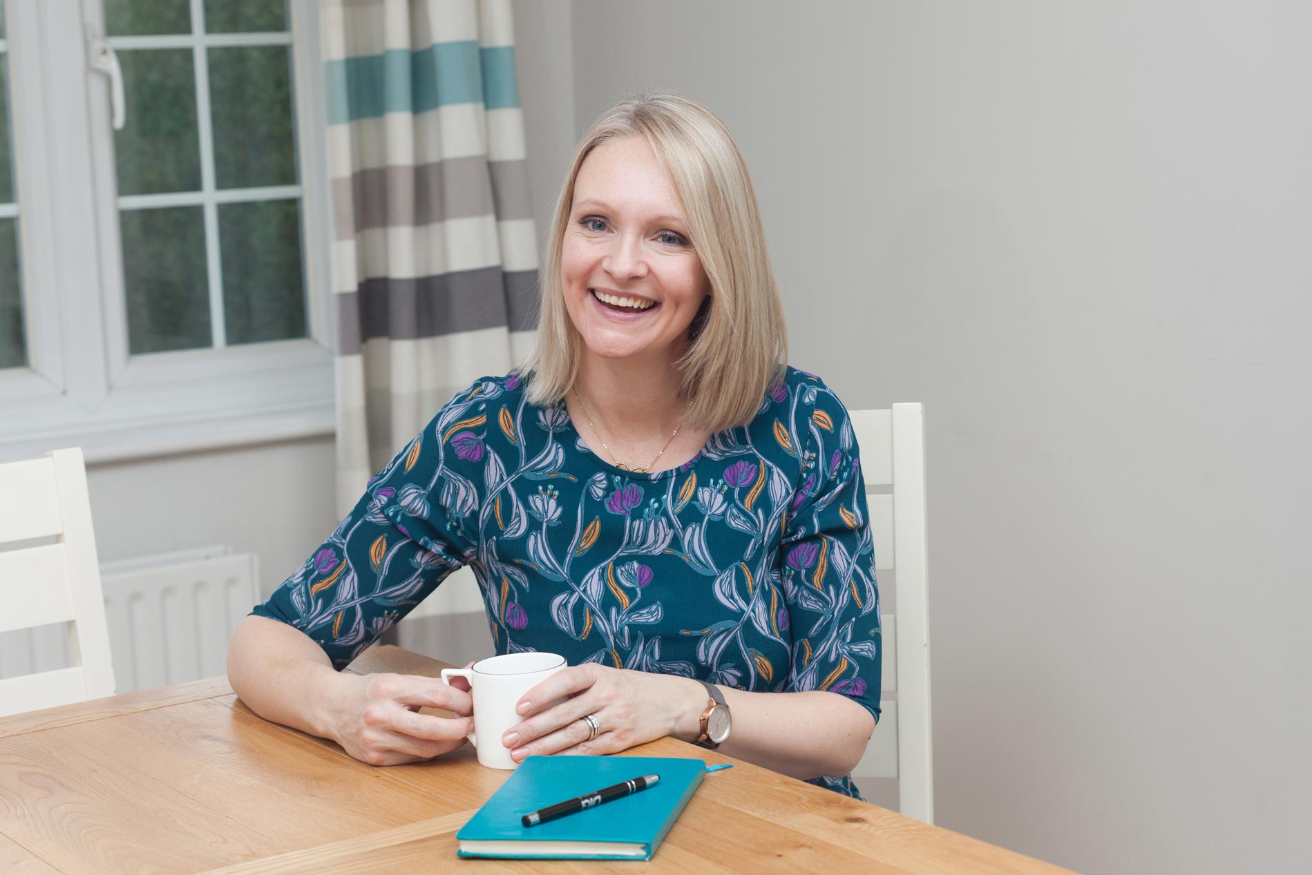 A smiling woman sits at a table with a cup of coffee. An example of personal branding photography in Croydon, Surrey & London.