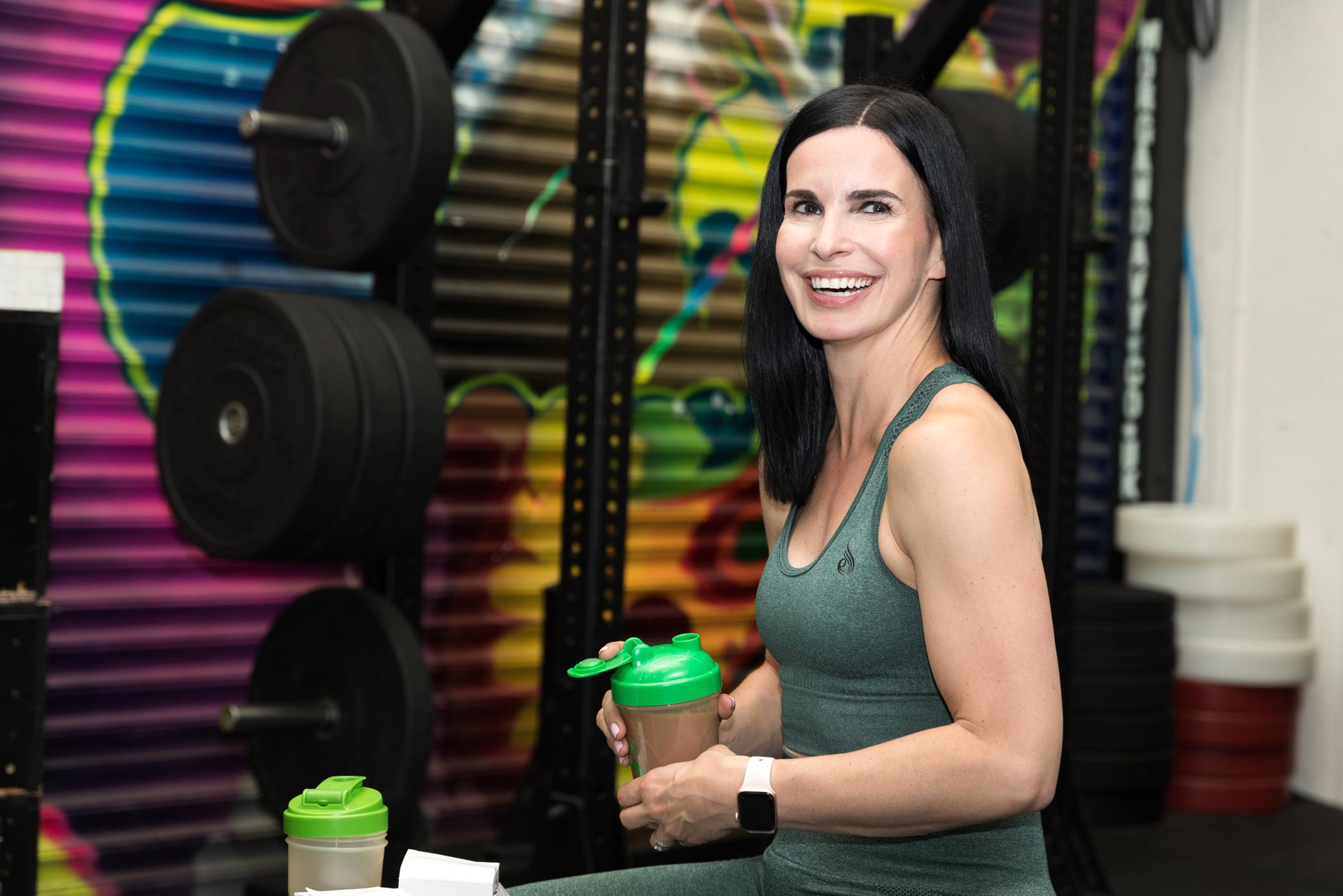 A smiling lady sitting in front of the weights rack at the gym during her fitness photography personal branding shoot in Croydon