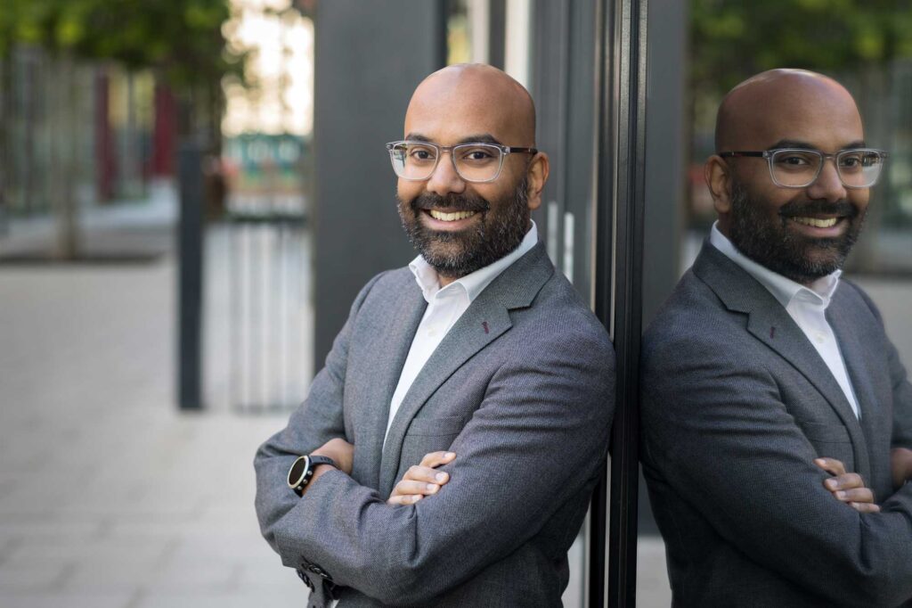 A business man in a suit posing for a headshot photo leans up against a glass building in Ruskin Square in Croydon