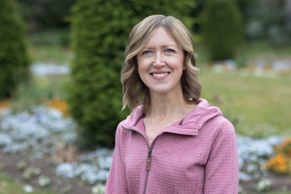 An example of what to wear for a professional headshot; a photo of a woman in a pink knit jumper taken outside with greenery as the background.