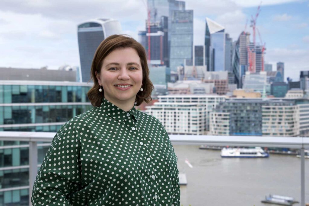 Professional London corporate headshot photography - a business woman wearing a green shirt with white spots, stands by the River Thames with the London city skyline in the background.