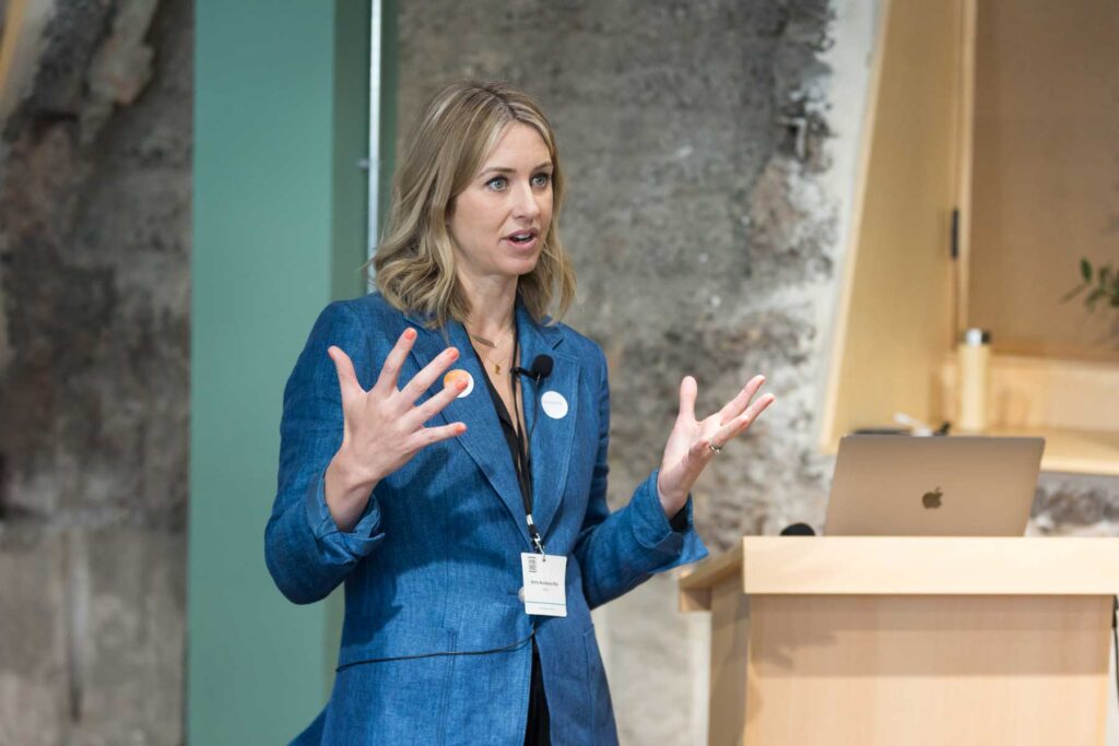 Photography showing a woman speaker at a London business event. She wears a blue jacket and stands with her hands in the air.