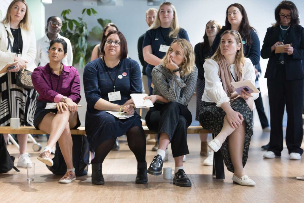 The audience listens to the speakers during a relaxed and informal corporate event in Central London. They sit in a row on a bench, listening intently.