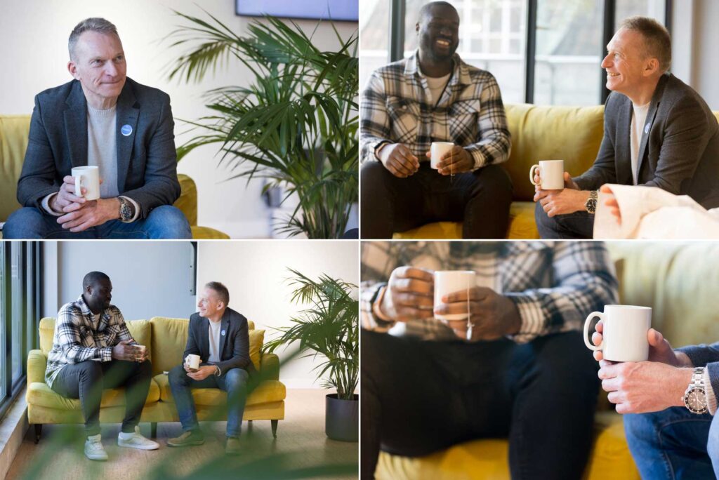 Two men sit on a dusky yellow sofa with cups of coffee having a conversation at the end of a business conference.